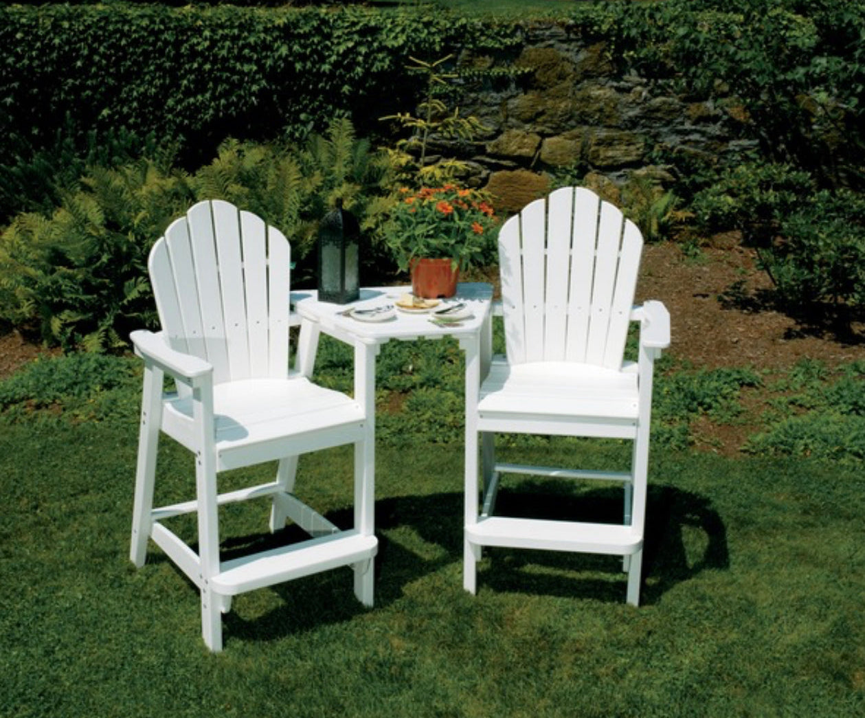 Two white Adirondack classic bar chairs with a table placed on grass against a backdrop of plants and a stone wall.